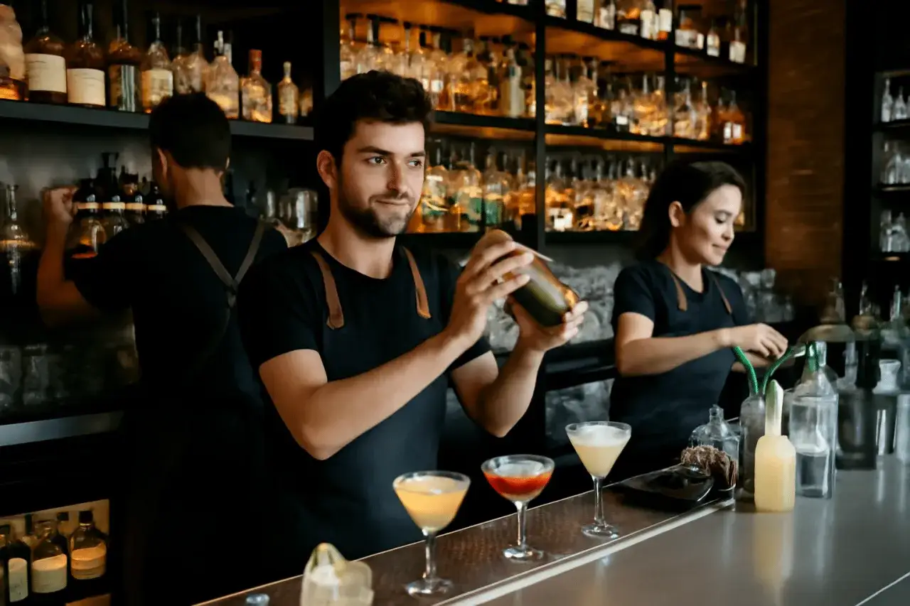 Barman preparando c&oacute;cteles con un shaker. Detr&aacute;s, otro empleado y una camarera ofrecen servicio en barra.