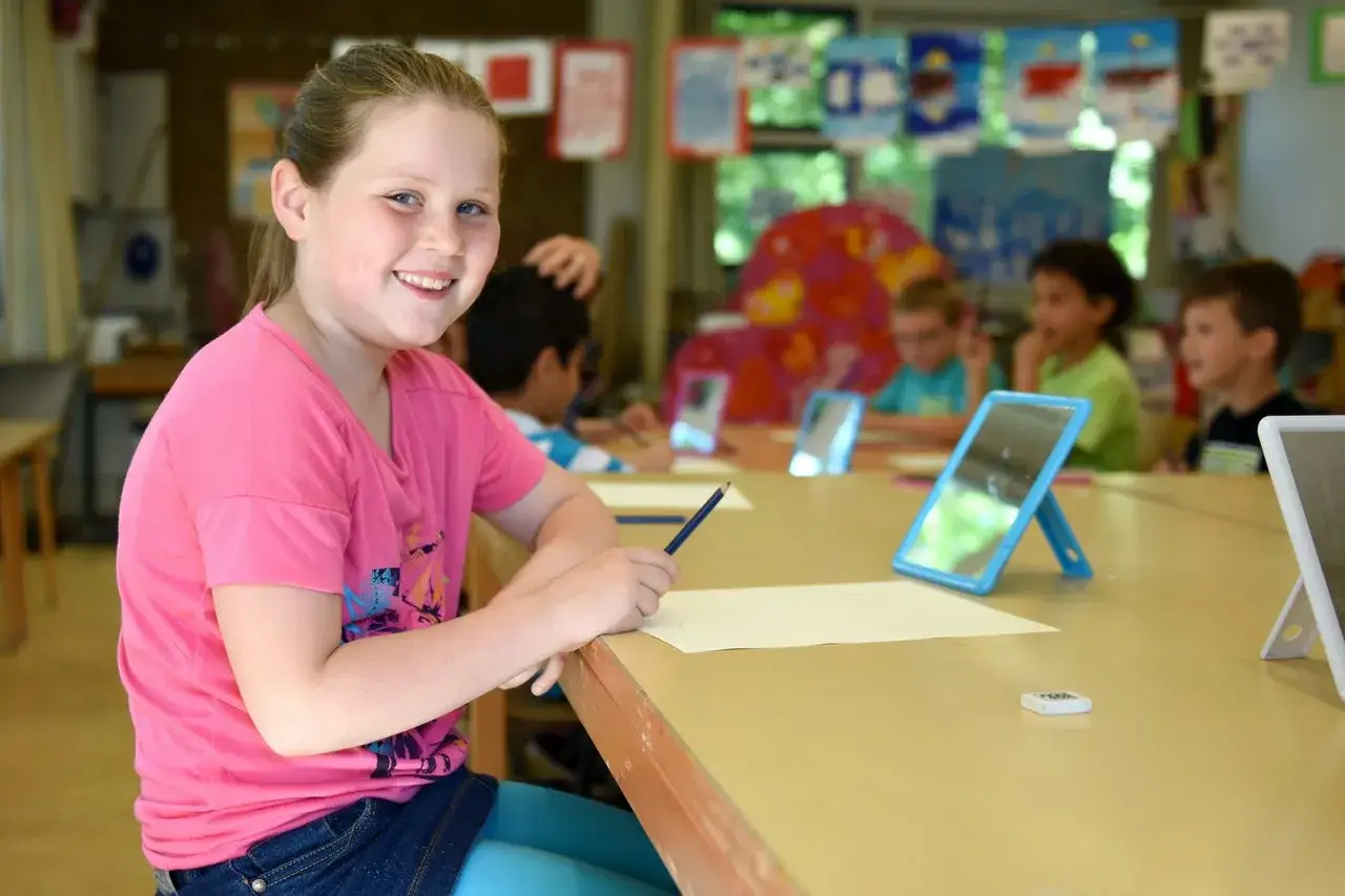 Een lachend meisje met een potlood in haar hand, omringd door klasgenoten. Dit is een voorbeeld van **didactisch coachen** in de klas.