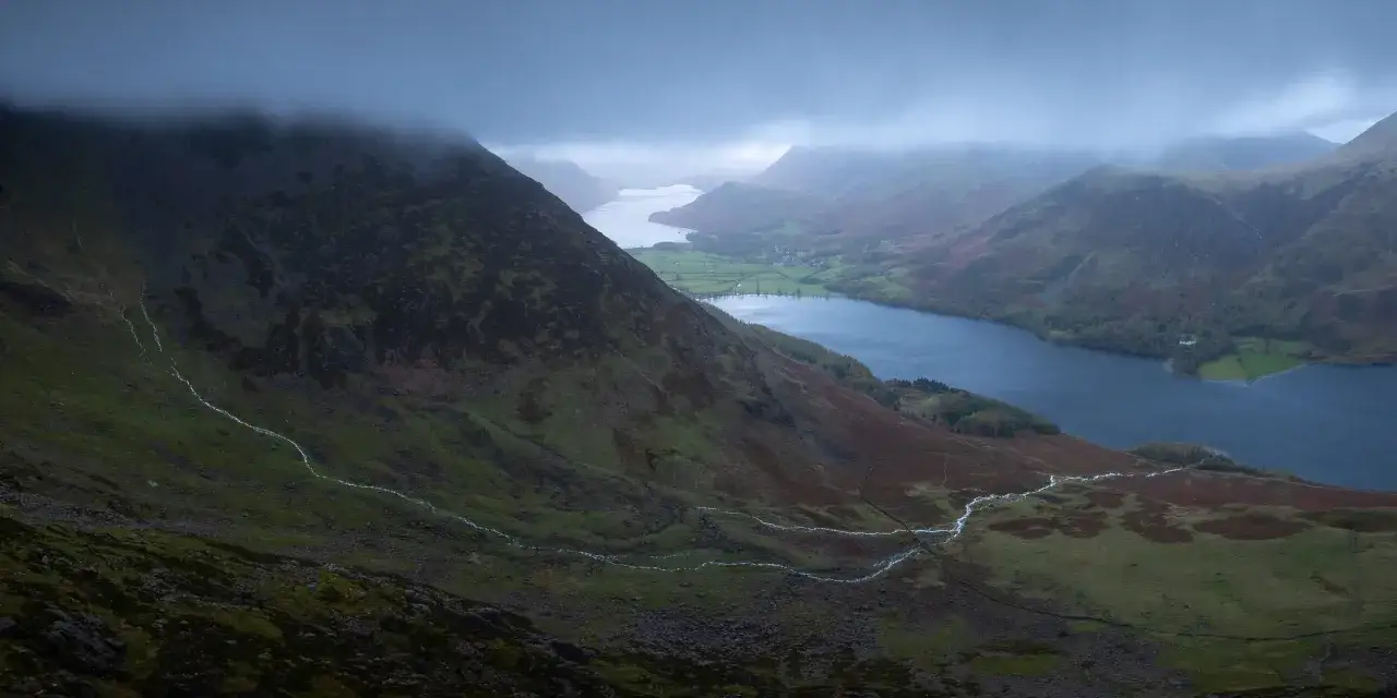 A dramatic aerial videography shot captures a misty mountain landscape with a winding river flowing into a dark blue lake.