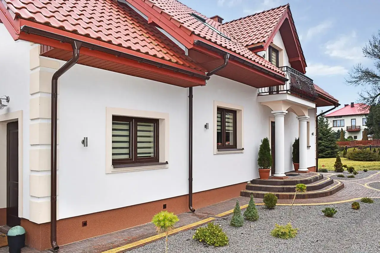 Elewacja domu z białym marmolitem, brązowymi oknami i czerwoną dachówką. Wokół zadbany ogród z kamyczkami i iglakami.
