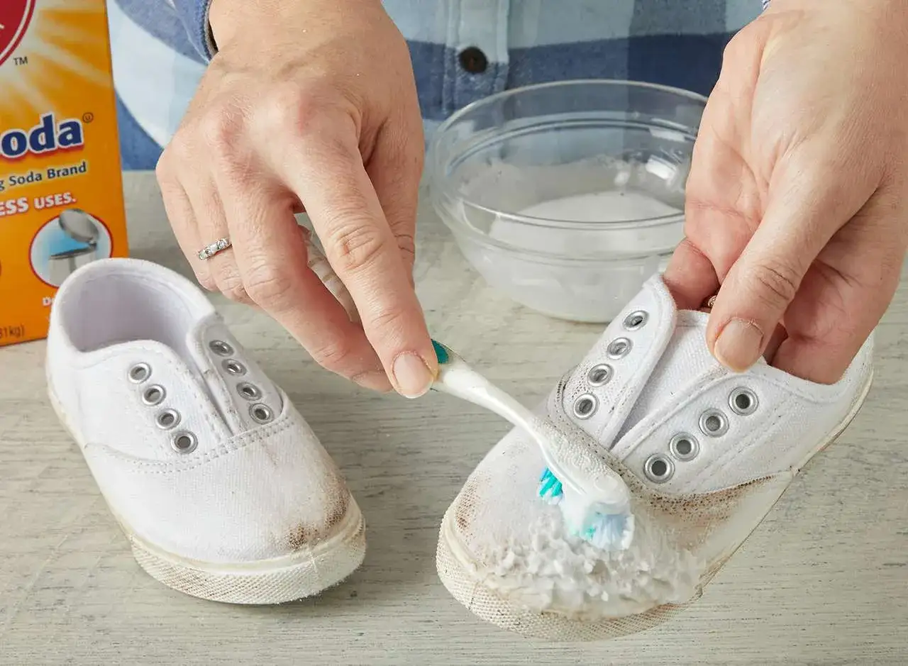 Manos limpian zapatillas blancas con cepillo y bicarbonato. Un taz&oacute;n con agua y una caja de bicarbonato de sodio completan la escena.