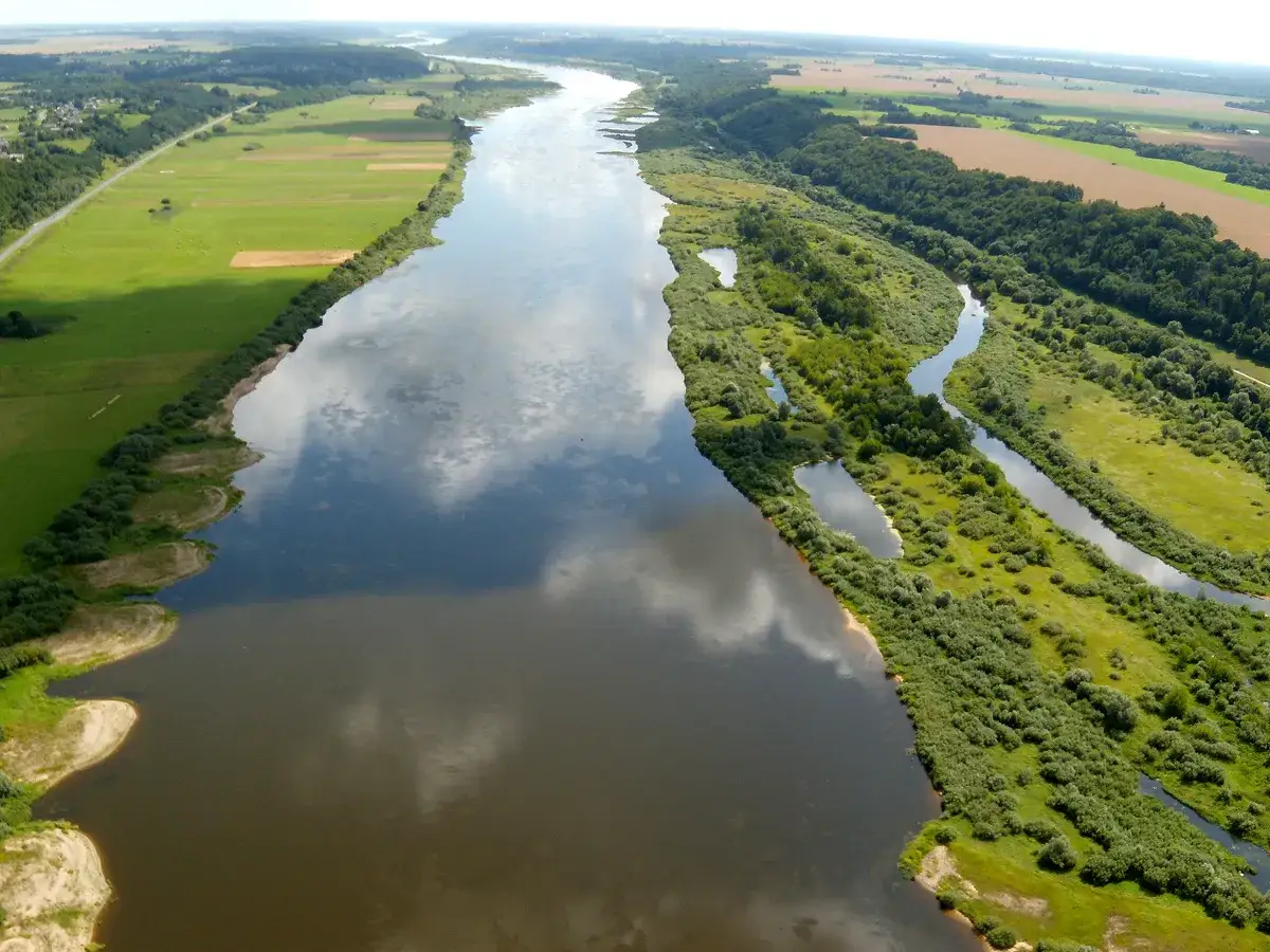 Szeroka rzeka Nemen meandruje przez zielone pola i lasy, odbijając chmury na swojej powierzchni.