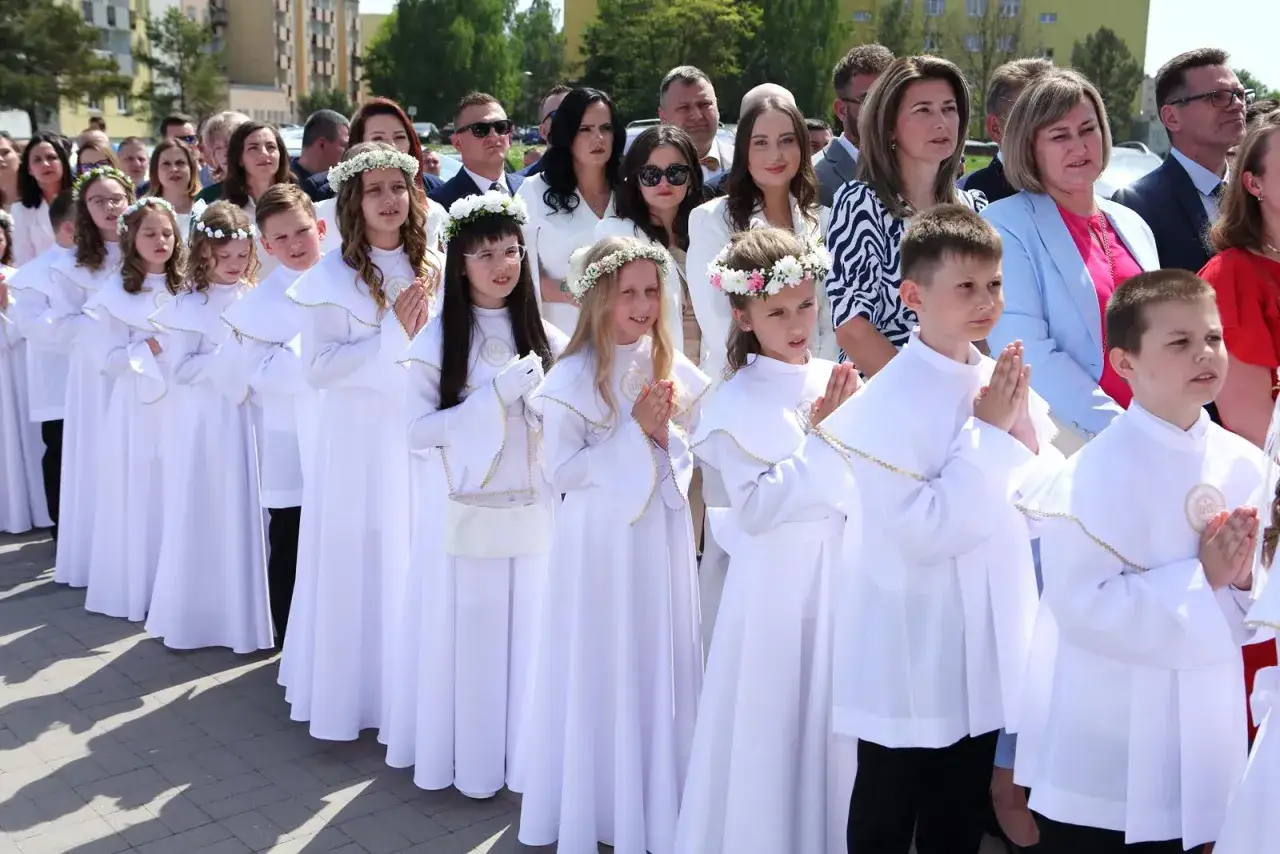 Dzieci w białych strojach, z wiankami na głowach, modlą się, gdy jest komunia. Rodzice stoją za nimi, obserwując uroczystość.