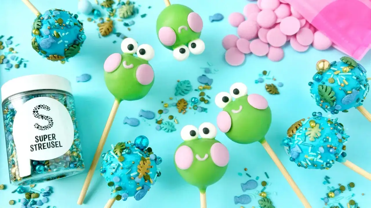 Süße Frosch-Cake-Pops und blaue Ozean-Cake-Pops, inspiriert von einem Cake Pops Rezept. Streusel und rosa Schokolade liegen bereit.