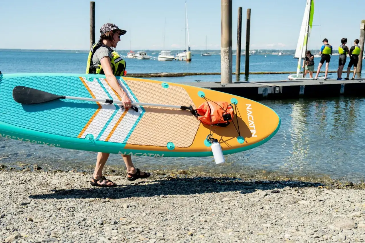 Donna con giubbotto salvagente porta una tavola sup colorata verso l'acqua. Sullo sfondo, bambini si preparano a salire su una barca a vela.