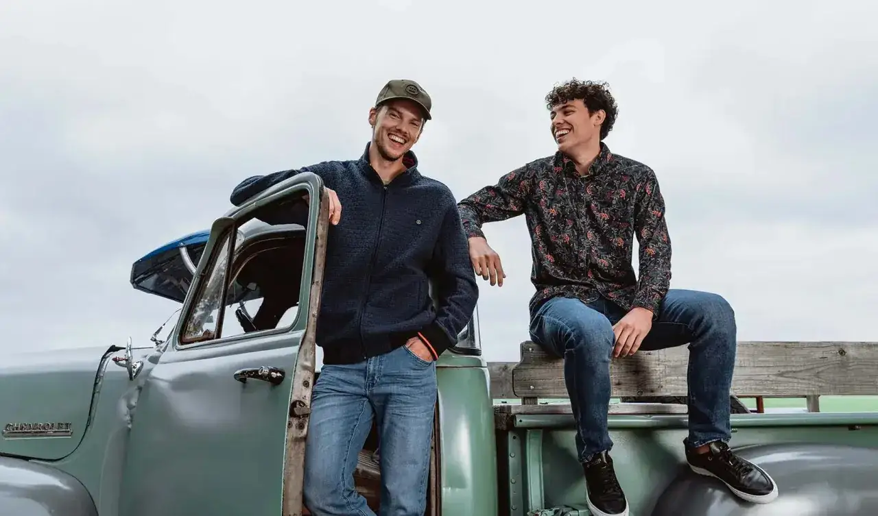 Zwei lachende M&auml;nner in l&auml;ssigen Jeans und Outfits lehnen an einem alten Pick-up.