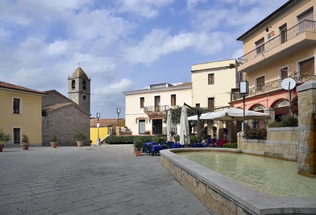Piazza con fontana e tavolini all'aperto nel **centro storico** di Arzachena. Campanile in pietra sullo sfondo.