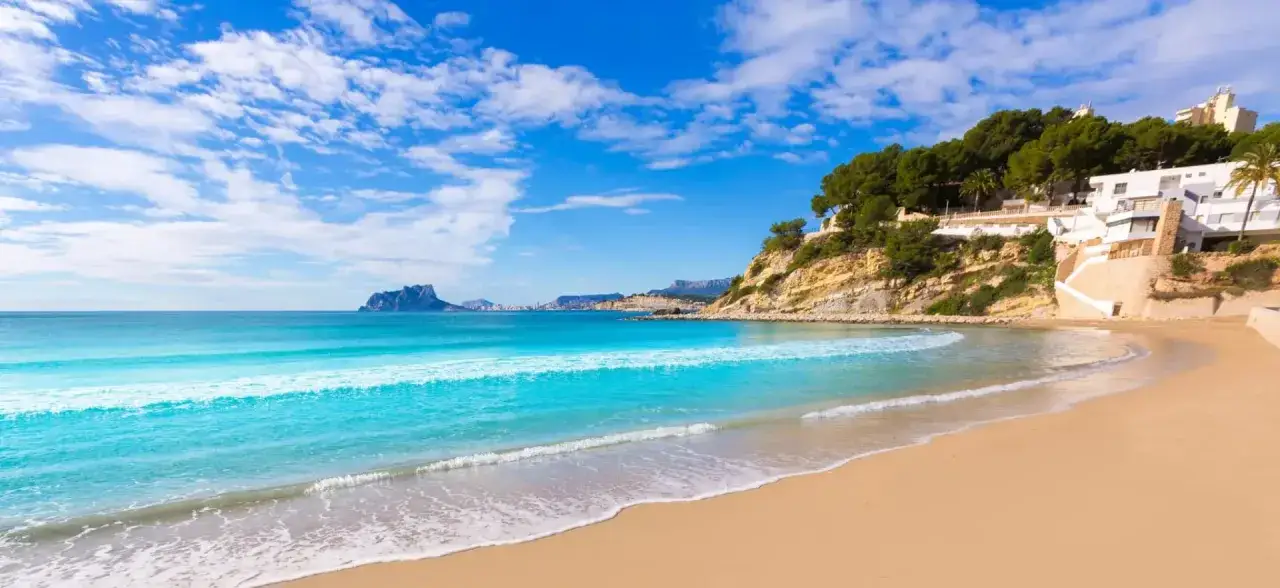 Playa de arena dorada y aguas turquesas, ideal para descubrir qué ver en Moraira. Edificios blancos coronan el acantilado verde.