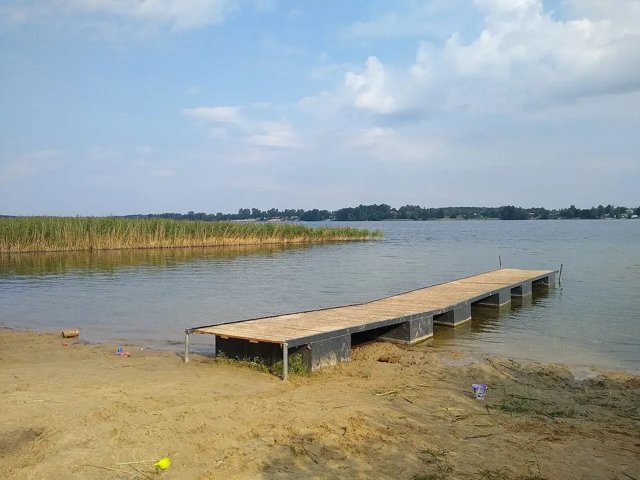 Drewniane molo na piaszczystym brzegu, trzciny i spokojne jezioro Białe Kaszuby pod błękitnym niebem.