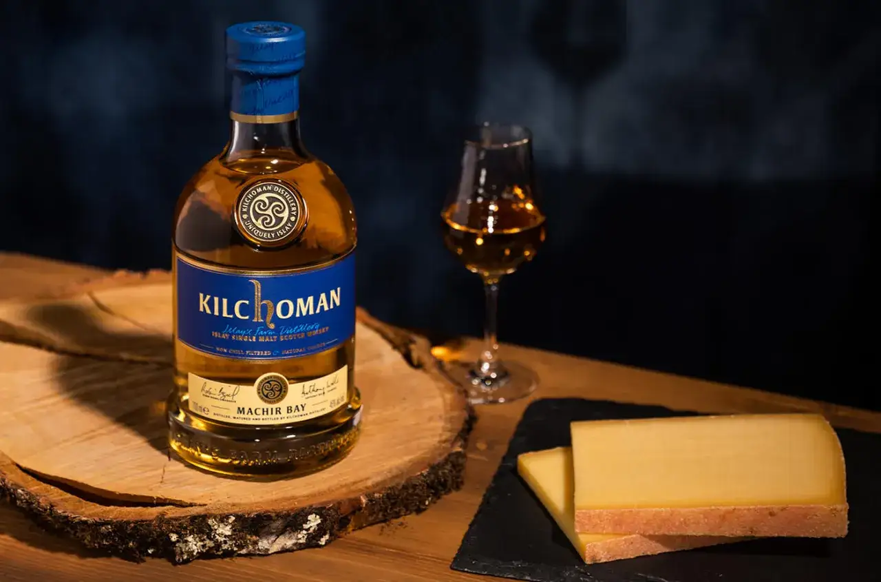 Bouteille de whisky Kilchoman Machir Bay et un verre de whisky, parfaits pour un digestif. Des tranches de fromage compl&egrave;tent la sc&egrave;ne.