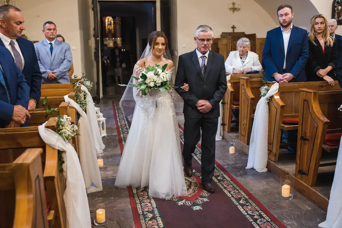 Panna młoda z bukietem idzie do ołtarza, prowadzona przez ojca. W kościele trwa ślub jednostronny, przysięga wkrótce zostanie złożona.