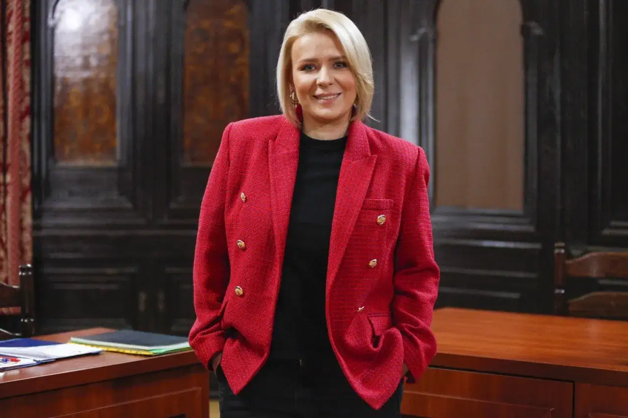 Uśmiechnięta blondynka w czerwonej marynarce, stojąca przy biurku. Ile lat ma Aneta Zając? Tego zdjęcie nie zdradza, ale wygląda na pewną siebie.
