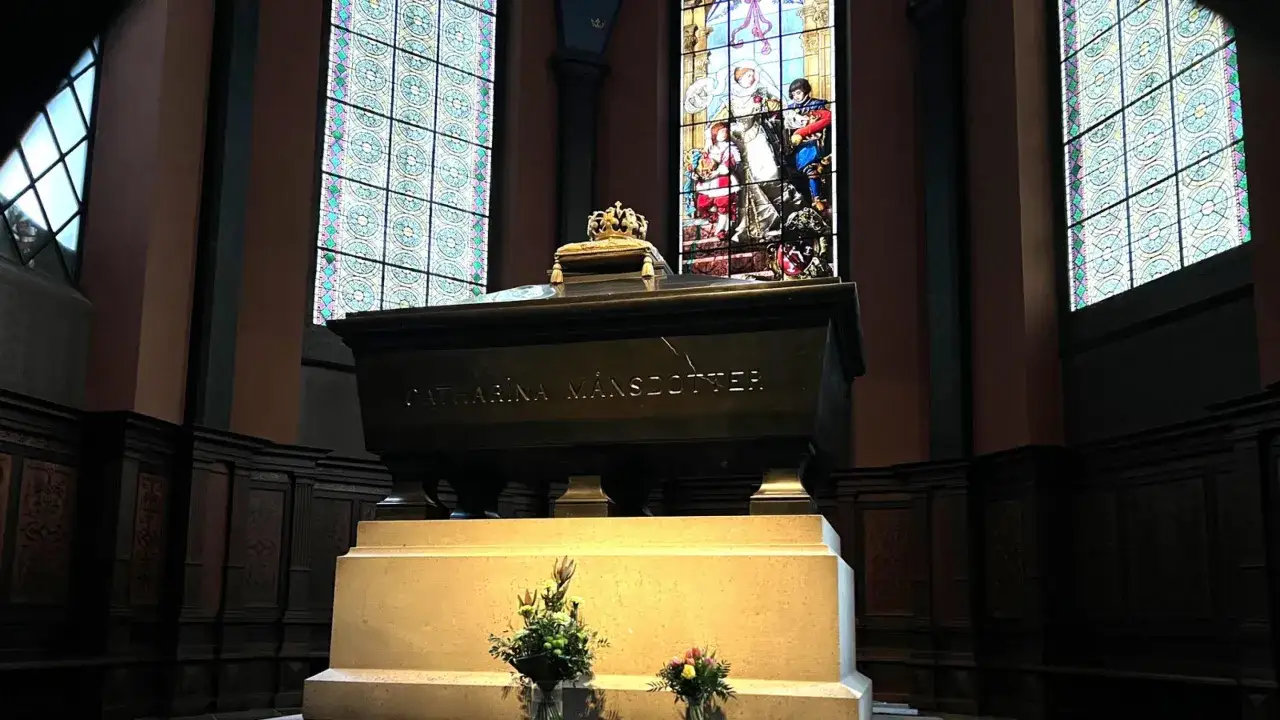 Gravkammare med inskriptionen CATHARINA M&Aring;NSDOTTER. En krona vilar p&aring; sarkofagen. Bakom syns ett glasm&aring;lat f&ouml;nster med en kunglig familj.