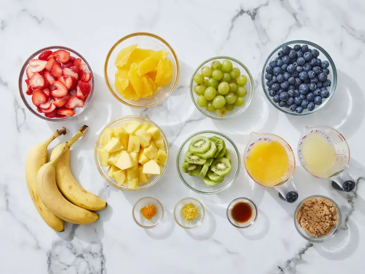 Zutaten für ein frisches obstsalat rezept: Erdbeeren, Orangen, Ananas, Bananen, Kiwis, Trauben, Blaubeeren, Orangensaft, Zitronensaft, brauner Zucker, Zimt und Zitronenschale.