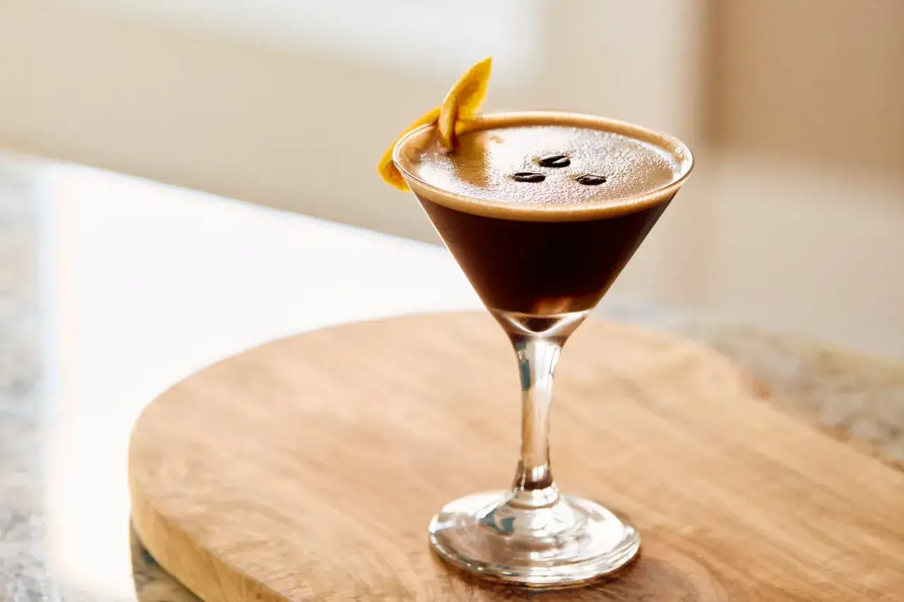 Elegancki coffee martini z pianką, trzema ziarnami kawy i sk&oacute;rką pomarańczy na drewnianej desce.