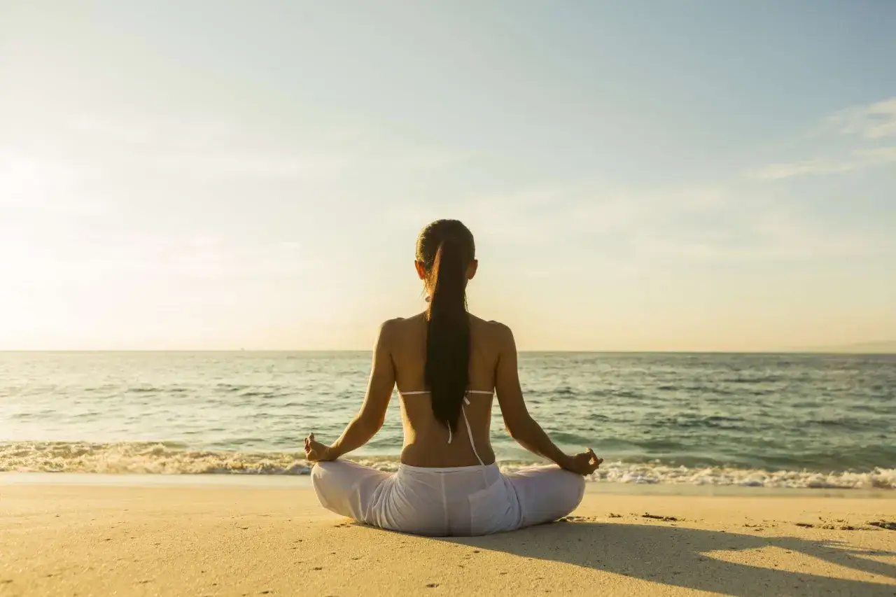 Frau in Lotusposition am Strand, die Meditation gegen Angst praktiziert. Sanfte Wellen und goldener Sonnenschein schaffen eine friedliche Atmosph&auml;re.