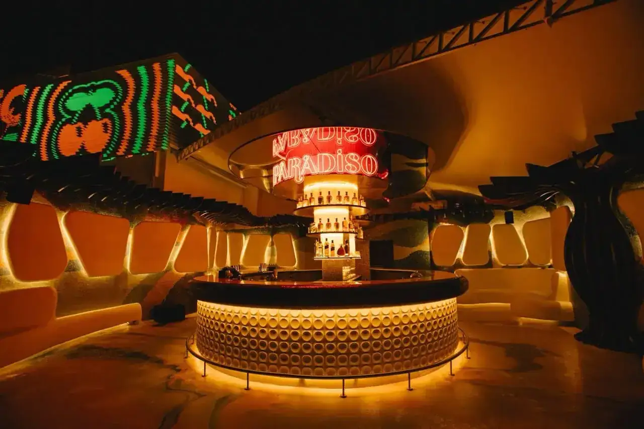 El interior de un club nocturno con un bar circular, luces de ne&oacute;n rojas que dicen 