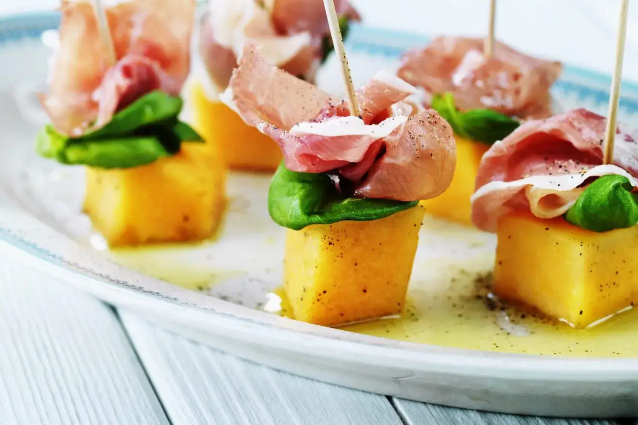 Sm&aring; t&auml;rningar av melon och skinka p&aring; spett, dekorerade med gr&ouml;na blad och peppar. En klassisk och l&auml;cker kombination.