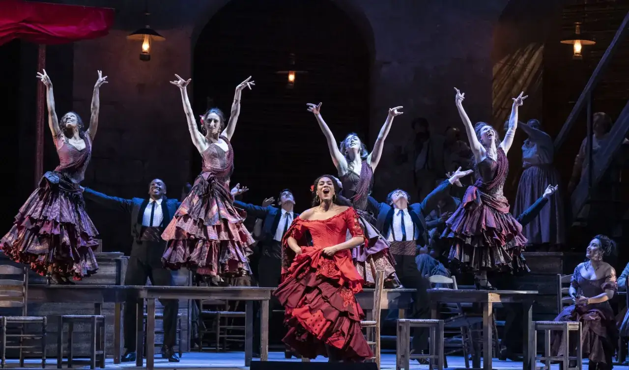 Carmen w czerwonej sukni śpiewa na scenie, otoczona tancerkami w hiszpańskich strojach i mężczyznami w garniturach.