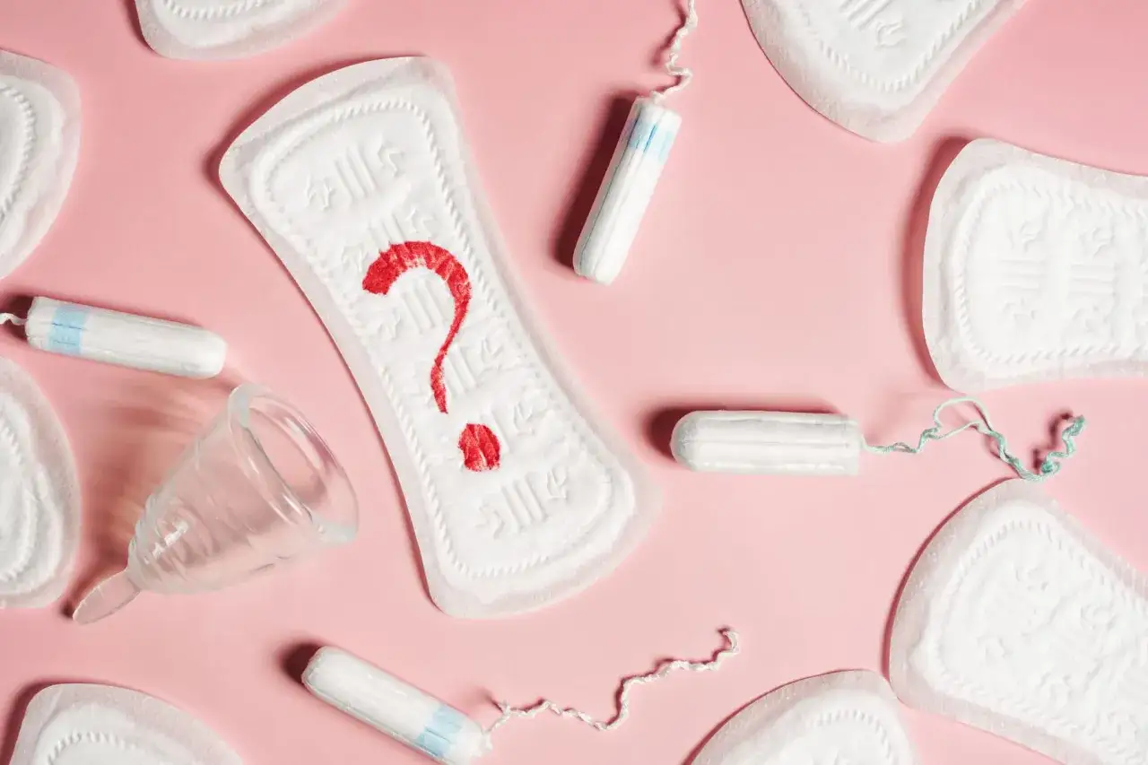 Brązowe plamienie zamiast okresu? Na różowym tle podpaski, tampony i kubeczek menstruacyjny. Czerwony znak zapytania na jednej z podpasek symbolizuje niepewność.