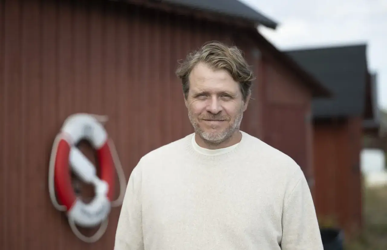 En man med blont h&aring;r och sk&auml;gg, som p&aring;minner om farbror Melkers &aring;lder, st&aring;r framf&ouml;r en r&ouml;d byggnad med en livboj.