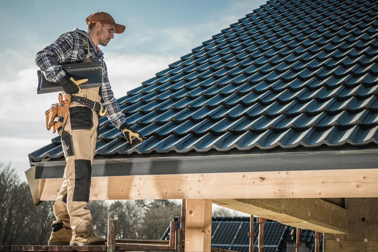 Un couvreur examine les mat&eacute;riaux de couverture bleus sur un toit en construction, sous un ciel clair.