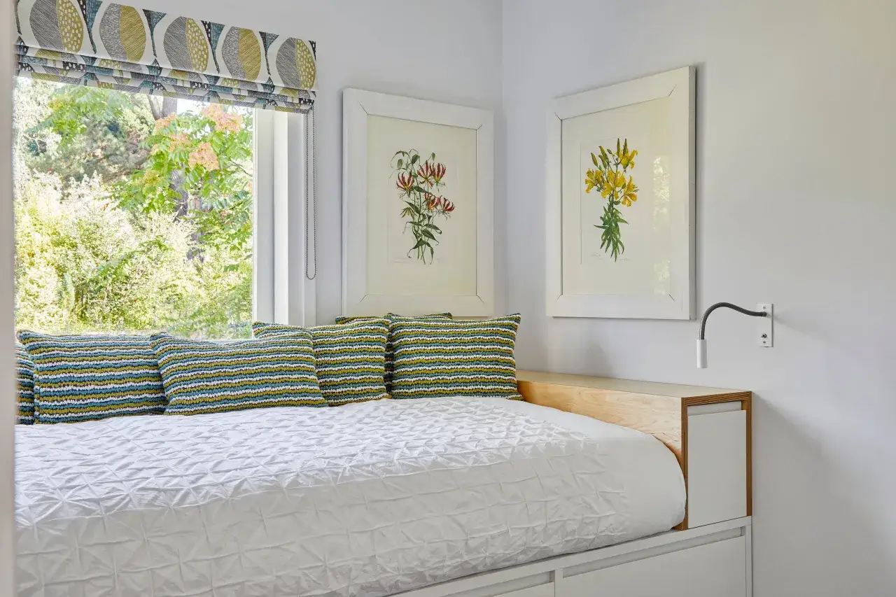 Cama en habitaci&oacute;n de 6 metros cuadrados, con cojines coloridos, cuadros de flores y ventana con vistas al jard&iacute;n.
