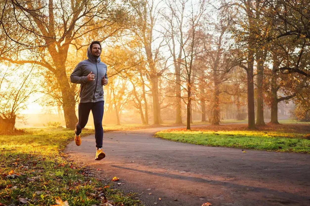 Uomo sorridente riprende a correre su un sentiero nel parco autunnale, illuminato dalla luce dorata del sole.