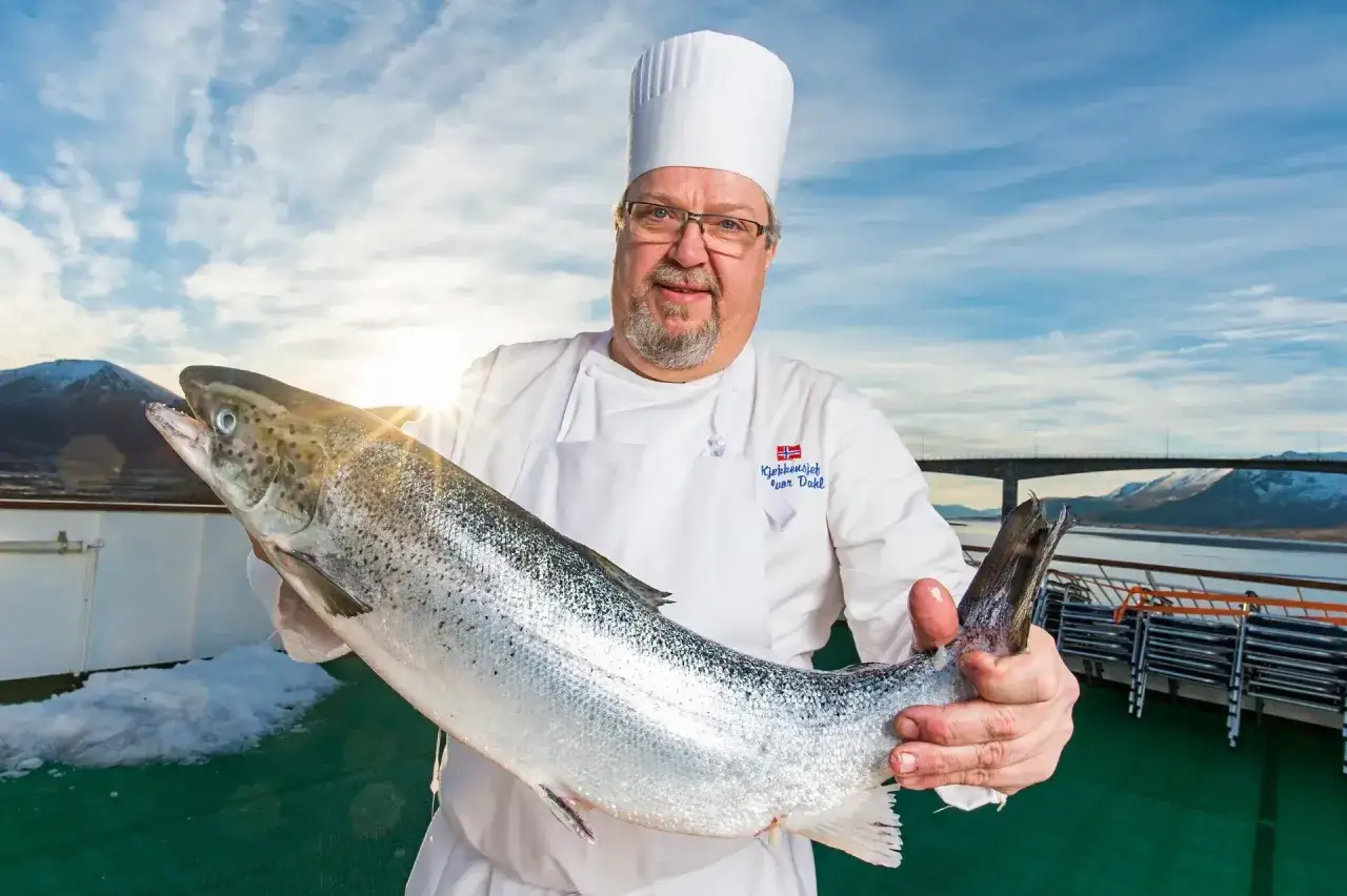 En kock i Norge stoltserar med en stor, f&auml;rsk fisk. Bakom skymtar en bro och sn&ouml;kl&auml;dda berg under en bl&aring; himmel.