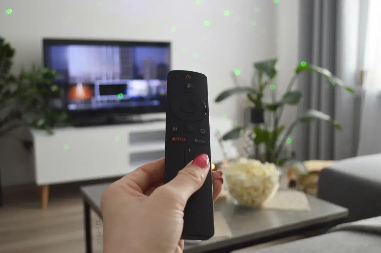 Dłoń trzyma pilot z przyciskiem Netflix, gotowa do wyboru polecane filmy Netflix. Obok miska popcornu.