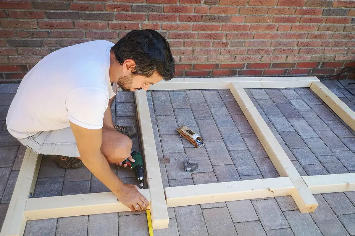 Un homme assemble un cache poubelle bois à faire soi-même. Il utilise une perceuse pour fixer les planches.