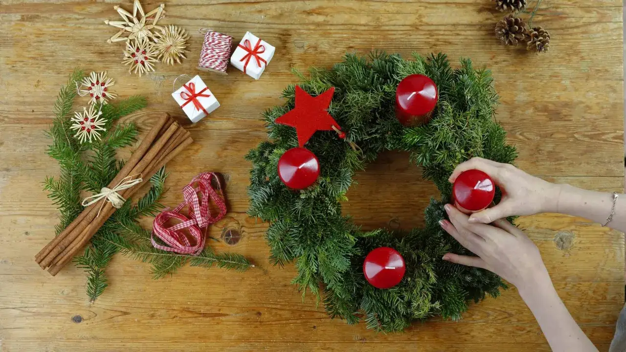 Hände schmücken einen Adventskranz mit roten Kerzen. Zimtstangen, Strohsterne und Geschenke erinnern an die Geschichte von Weihnachten.