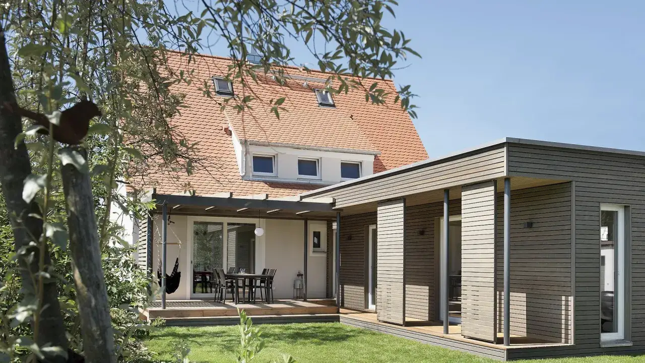 Moderner Anbau an altes Haus mit Holzfassade und Terrasse. Ein Schaukelstuhl h&auml;ngt unter dem Vordach.