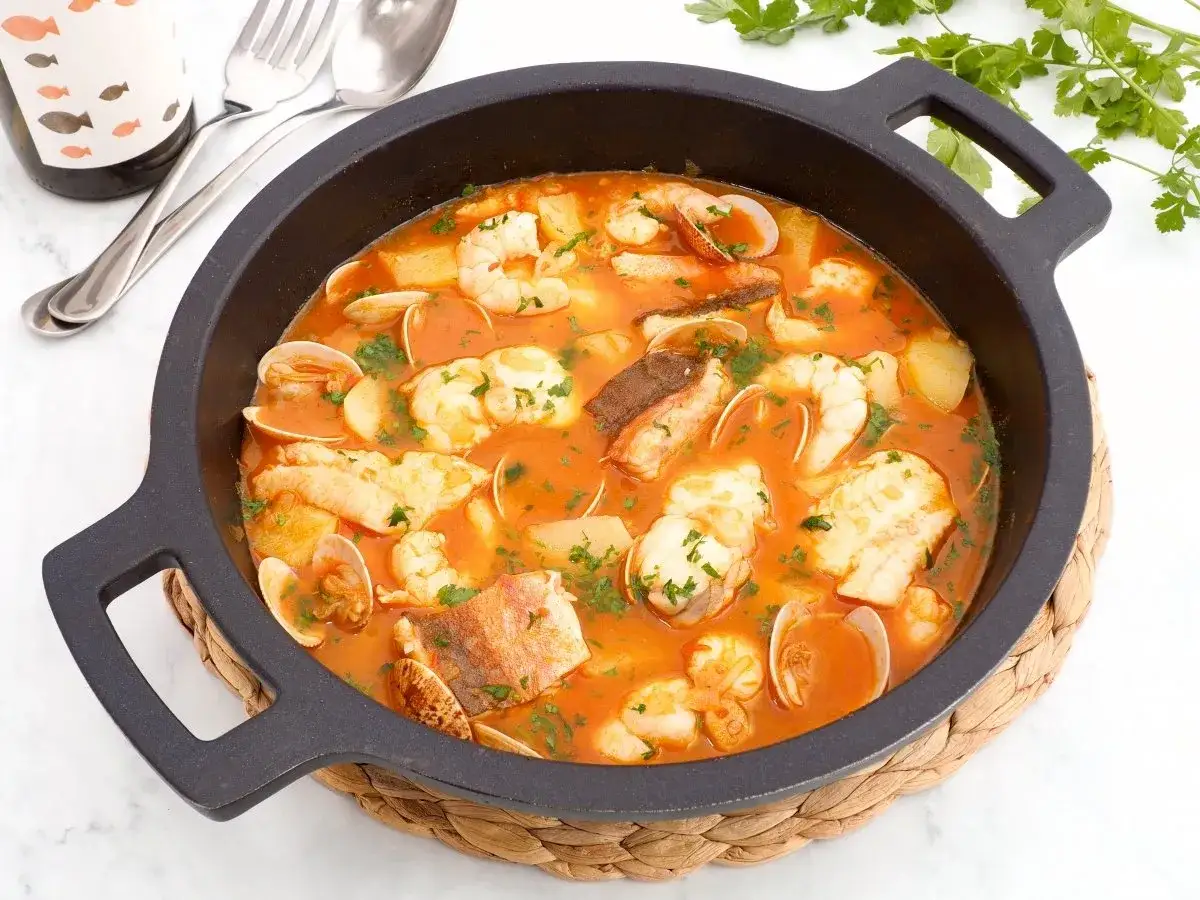 Caldereta de pescado humeante, con trozos de pescado blanco, gambas y almejas en una rica salsa roja, decorada con perejil fresco.