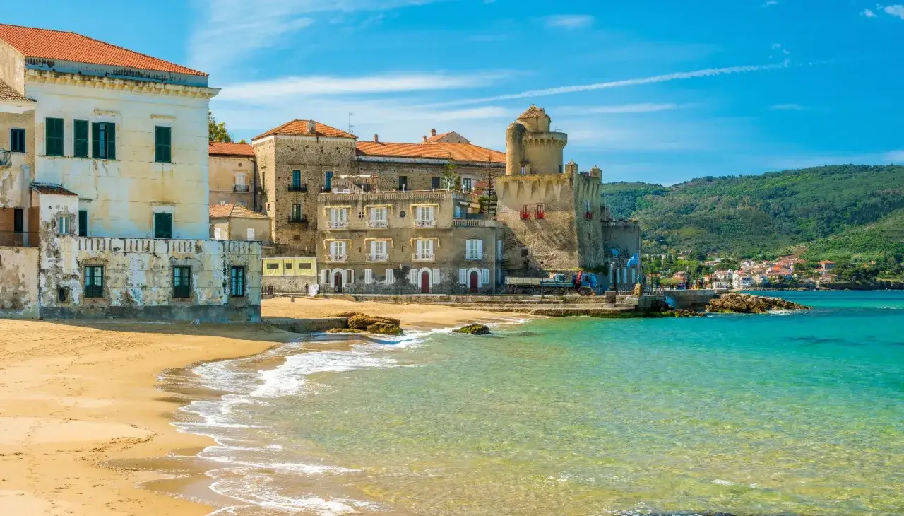Edifici storici e un castello si affacciano su una spiaggia dorata e acque cristalline, un incantevole scorcio della costiera cilentana.