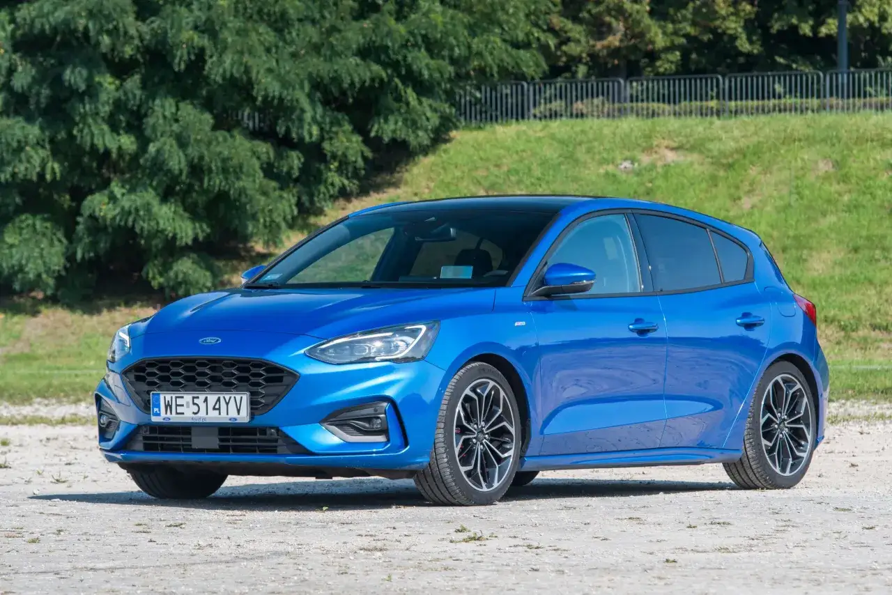 Błękitny Ford Focus 1.0 EcoBoost z czarnym dachem i sportowymi felgami stoi na żwirowej nawierzchni, na tle zieleni.