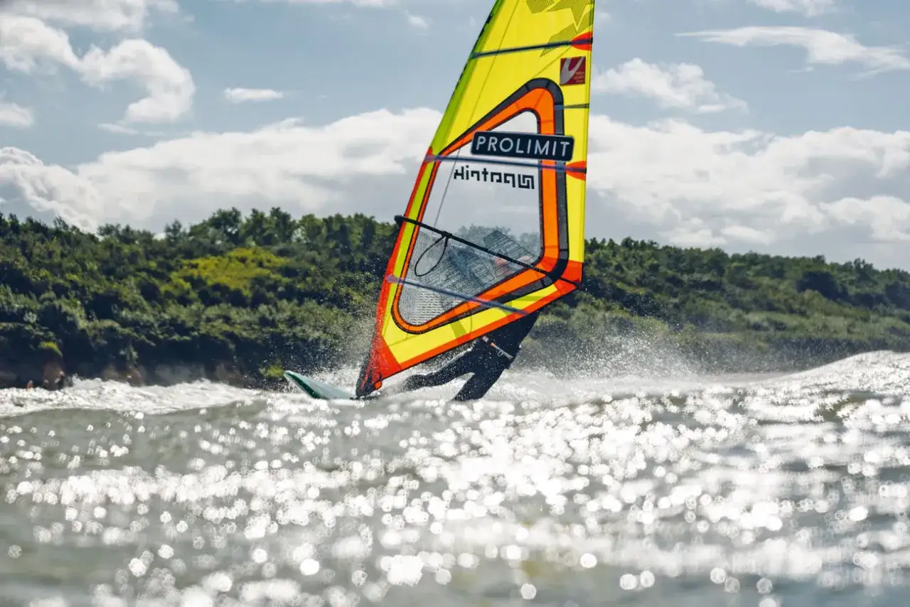 Windsurfer gleitet über die Ostsee, die Sonne funkelt auf den Wellen. Ein bunter Segel dominiert das Bild.
