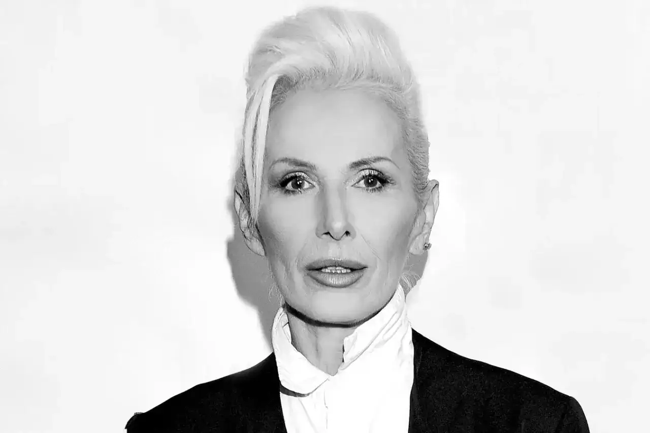 Elegancka kobieta z siwymi włosami, w czarnej marynarce i białej koszuli. Jej kora wiek dodaje jej charyzmy.