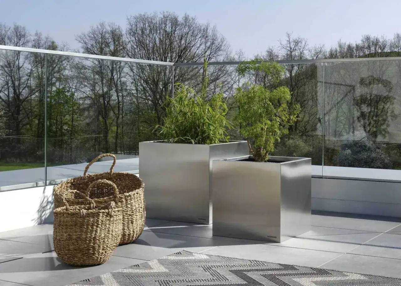 Moderne rechteckige Pflanzkübel aus Edelstahl mit grünen Pflanzen, ideal zum **dachterrasse bepflanzen**. Daneben zwei geflochtene Körbe.