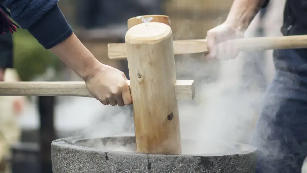 H&auml;nde schlagen mit Holzst&ouml;&szlig;eln auf ged&auml;mpften Reis in einem Steinm&ouml;rser. Ein traditionelles mochi rezept wird hier lebendig.