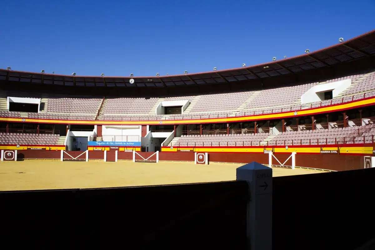 Interior de la plaza de toros de Roquetas de Mar, con gradas vacías y el ruedo de arena bajo un cielo azul.