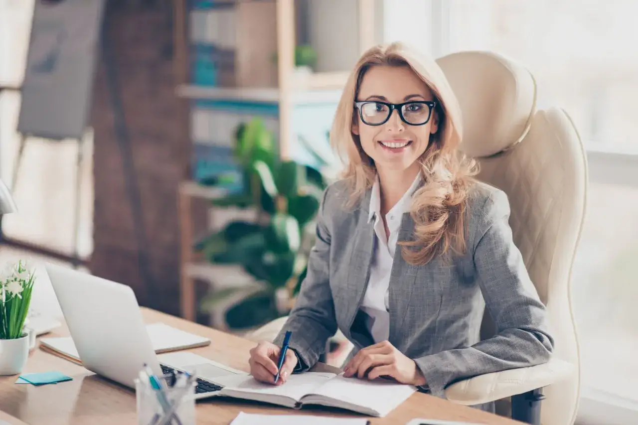 Donna in ufficio, con occhiali e tailleur grigio, scrive su un quaderno. Potrebbe occuparsi di una procura generale notarile.