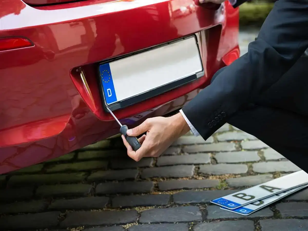 Un uomo in giacca e cravatta monta una targa su un'auto rossa, rispettando l'art. 102 CDS. Due targhe sono a terra.