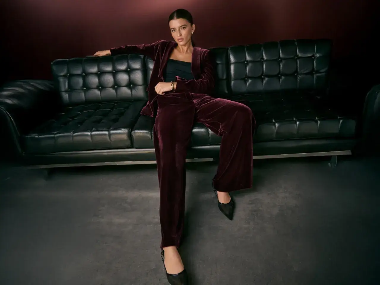 A woman in a burgundy velvet suit poses on a black leather couch, embodying the perfect christmas party dress code.
