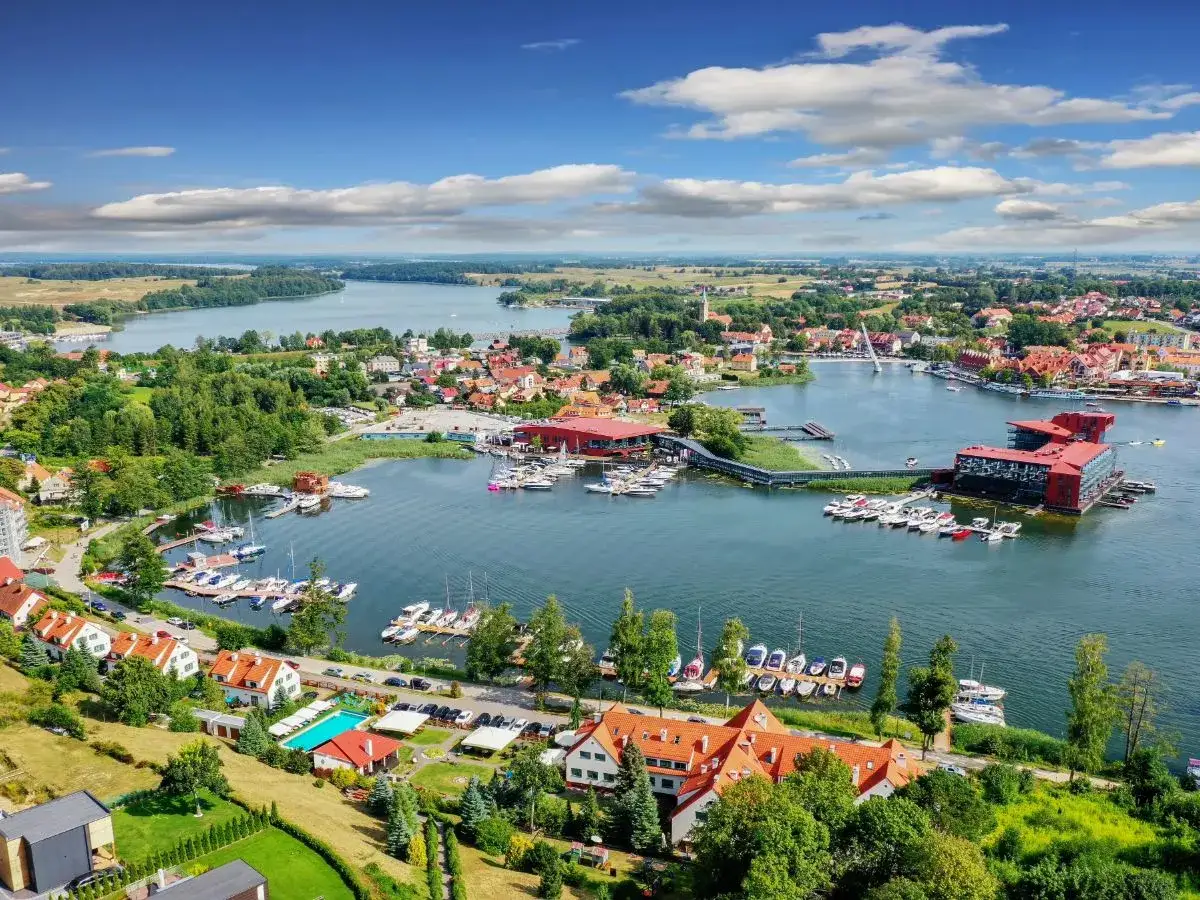 Malownicze Mazury: port z łodziami, czerwone dachy domów i nowoczesne budynki nad jeziorem. Atrakcje dla żeglarzy i miłośników wypoczynku.