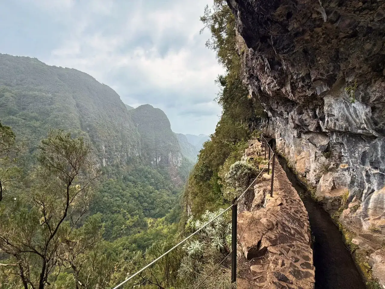 A narrow stone path hugs a cliff face along the Levada do Moinho, with a water channel beside it. Lush green valleys stretch out into the distance under a cloudy sky.