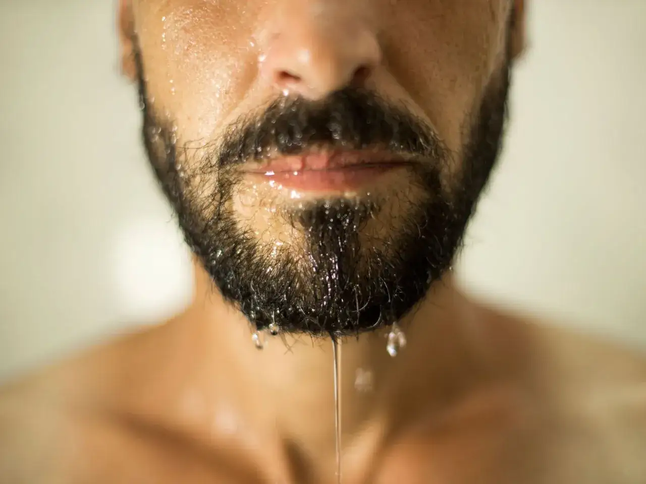 Primer plano de un hombre con barba oscura y húmeda, gotas de agua resbalan por su mentón. Las perillas de agua brillan.