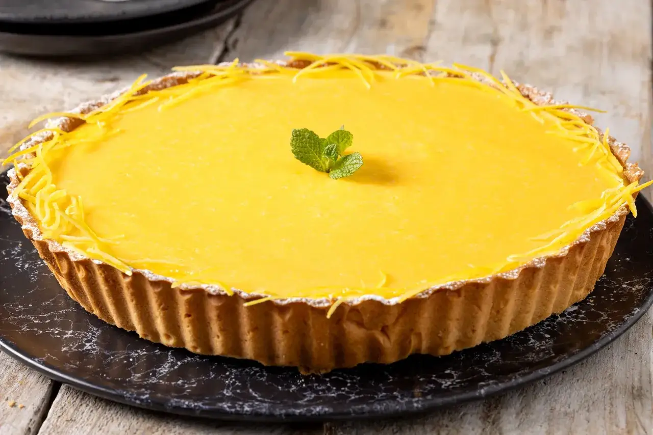 Una deliziosa crostata al limone, con scorzette gialle e una fogliolina di menta al centro, su un piatto nero.