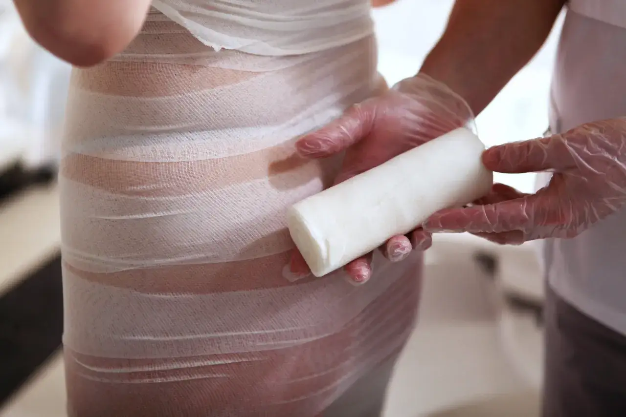 Dłonie w rękawiczkach trzymają rolkę bandaża, przygotowując się do zabiegu body wrapping. Widoczne są efekty zabiegu na ciele.