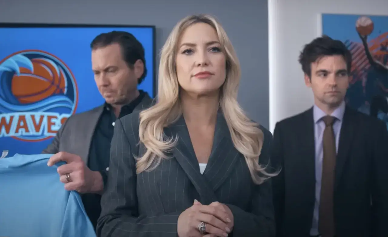 Kate Hudson och två män står framför en skärm med en basketboll och vågor. En man håller upp en ljusblå tröja. De verkar vara på en **running point** för ett sportlag.